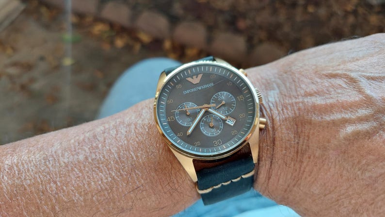 May include: Gold-toned wristwatch with a brown face and black leather band. The watch features multiple subdials and the brand name "Emporio Armani" is visible on the face. The watch is secured on a wrist.