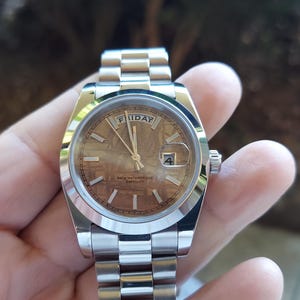 May include: A silver wristwatch with a brown face, displaying the day as "FRIDAY" and the date as "4". The watch has a silver band and a small crown. The watch is held in a hand.