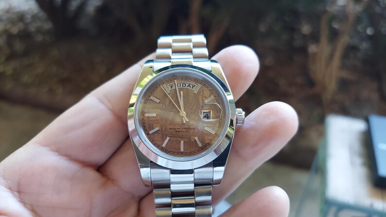 May include: A silver wristwatch with a brown face, displaying the day as "FRIDAY" and the date as "4". The watch has a silver band and a small crown. The watch is held in a hand.