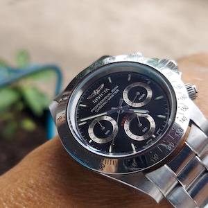 May include: A stainless steel Invicta Professional Chrono-Master watch with a black dial. The watch features three sub-dials and a silver bezel with white markings. The watch is on a stainless steel strap.