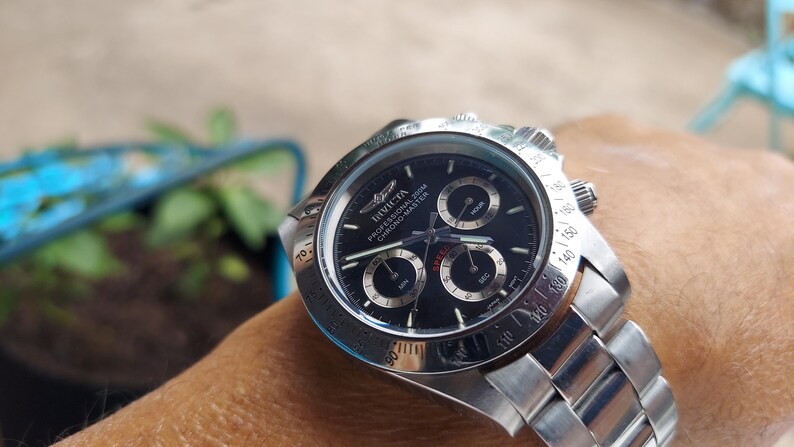 May include: A stainless steel Invicta Professional Chrono-Master watch with a black dial. The watch features three sub-dials and a silver bezel with white markings. The watch is on a stainless steel strap.