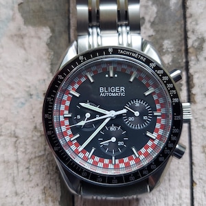 May include: A stainless steel Bliger Automatic watch with a black face, white hands, and red and white accents. The watch features a tachymeter bezel and three sub-dials. The watch is on a stainless steel band.