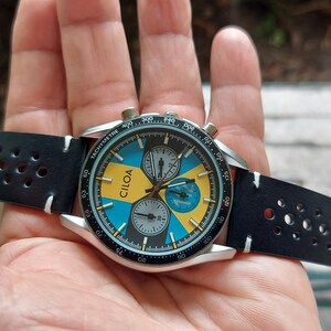 Pu&ograve; includere: Un orologio cronografo CILOA con cassa rotonda color argento, lunetta tachimetrica nera e quadrante con segmenti alternati blu e gialli. L'orologio include due sotto-quadranti e un cinturino in pelle nera traforata con cuciture bianche.