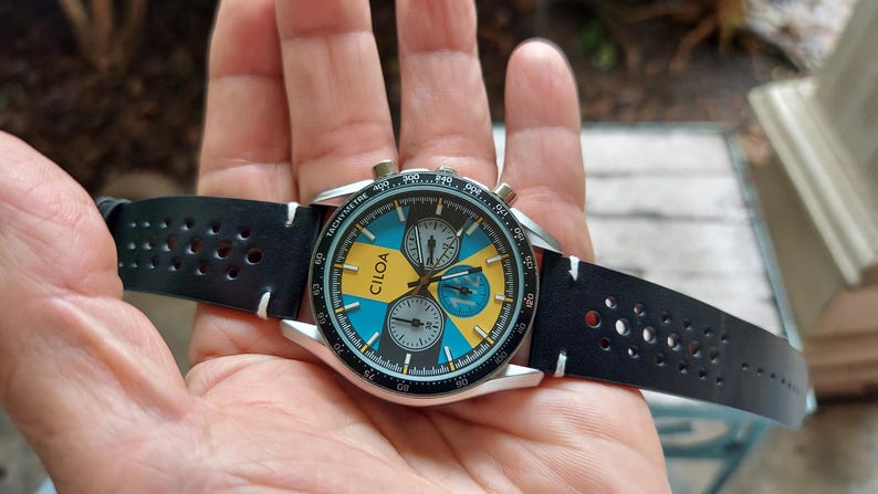 Pu&ograve; includere: Un orologio cronografo CILOA con cassa rotonda color argento, lunetta tachimetrica nera e quadrante con segmenti alternati blu e gialli. L'orologio include due sotto-quadranti e un cinturino in pelle nera traforata con cuciture bianche.