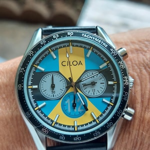 Pu&ograve; includere: Un orologio cronografo CILOA con un quadrante vivace caratterizzato da un motivo geometrico giallo e azzurro, sottoquadranti neri e una scala tachimetrica sulla lunetta esterna. Il quadrante mostra il numero 112 in un accento circolare blu.