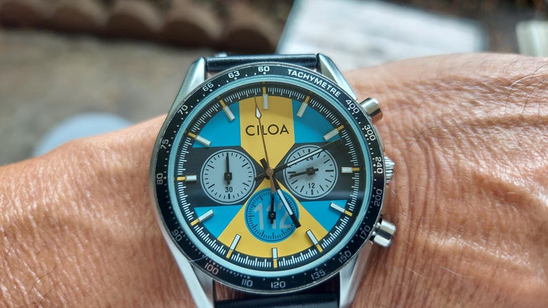 Pu&ograve; includere: Un orologio cronografo CILOA con un quadrante vivace caratterizzato da un motivo geometrico giallo e azzurro, sottoquadranti neri e una scala tachimetrica sulla lunetta esterna. Il quadrante mostra il numero 112 in un accento circolare blu.