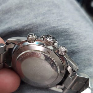 May include: A silver stainless steel wristwatch with a metal strap. The watch has a round face with three small dials and three buttons. The back of the watch is visible, with the words "STAINLESS STEEL" and "20ATM" engraved.