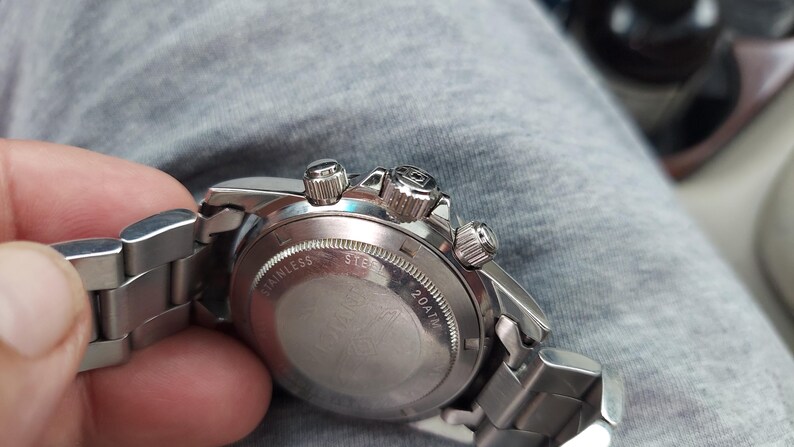 May include: A silver stainless steel wristwatch with a metal strap. The watch has a round face with three small dials and three buttons. The back of the watch is visible, with the words "STAINLESS STEEL" and "20ATM" engraved.