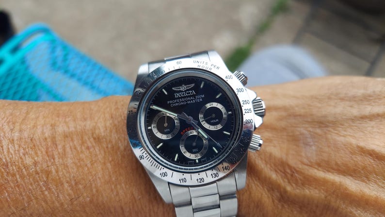 May include: A stainless steel Invicta Chrono-Master watch with a black dial. The watch has three sub-dials, a date display, and a tachymeter bezel. The watch is on a stainless steel bracelet.