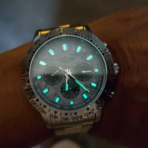 May include: A silver wristwatch with a dark gray face and luminous green markers and hands. The watch has three sub-dials and a rotating bezel with white numerals. The watch is on a silver band.