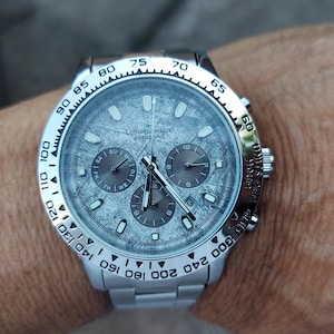 May include: A silver wristwatch with a textured gray face and three sub-dials. The watch has a silver bezel with black markings and a silver band. The watch is on a wrist.