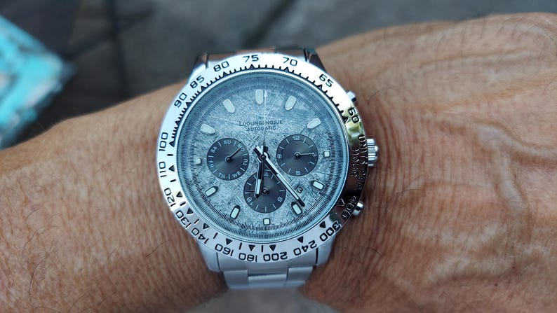 May include: A silver wristwatch with a textured gray face and three sub-dials. The watch has a silver bezel with black markings and a silver band. The watch is on a wrist.