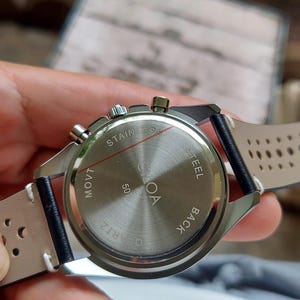 Pu&ograve; includere: Un fondello per orologio in acciaio inossidabile con finitura spazzolata circolare, recante la scritta "STAINLESS STEEL BACK" e "MOVT 50". L'orologio presenta un cinturino in pelle nera con dettagli traforati e cuciture bianche e tre pulsanti.