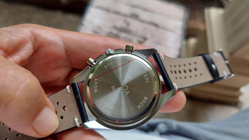 Pu&ograve; includere: Un fondello per orologio in acciaio inossidabile con finitura spazzolata circolare, recante la scritta "STAINLESS STEEL BACK" e "MOVT 50". L'orologio presenta un cinturino in pelle nera con dettagli traforati e cuciture bianche e tre pulsanti.