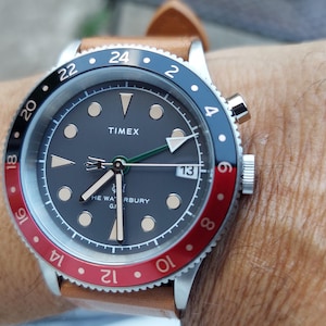 May include: A Timex Waterbury GMT wristwatch with a dark gray dial, luminous hour markers, and a date window at the three o'clock position. The watch features a two-tone red and black rotating bezel with 24-hour markings and a tan leather strap.