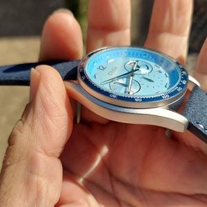May include: A blue and silver wristwatch with a light blue face and a blue leather strap. The watch has a silver case and a dark blue bezel. The watch face has three sub-dials and silver hands.