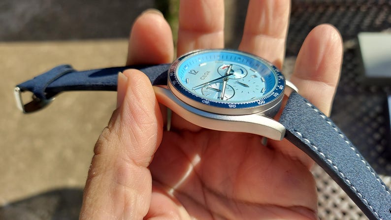 May include: A blue and silver wristwatch with a light blue face and a blue leather strap. The watch has a silver case and a dark blue bezel. The watch face has three sub-dials and silver hands.