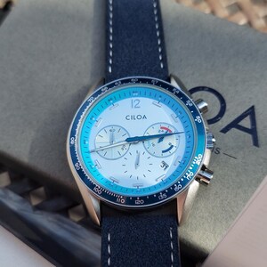 May include: A Ciloa watch with a blue and silver face and a navy blue leather strap. The watch has a silver case and a blue bezel. The watch is on top of a gray box with the Ciloa logo. The watch face has the number 12.