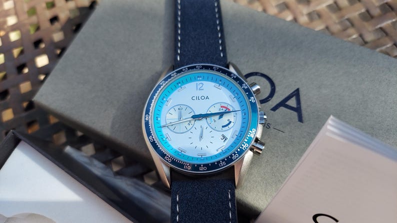 May include: A Ciloa watch with a blue and silver face and a navy blue leather strap. The watch has a silver case and a blue bezel. The watch is on top of a gray box with the Ciloa logo. The watch face has the number 12.