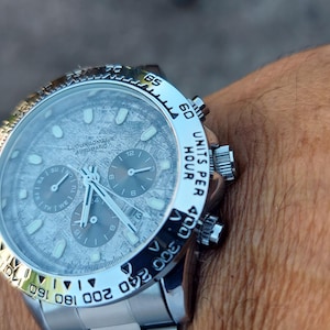 May include: A silver wristwatch with a textured gray face and three sub-dials. The watch has a silver bezel with black markings and a silver band. The watch face has the text "AUTOMATIC" and "UNITS PER HOUR".