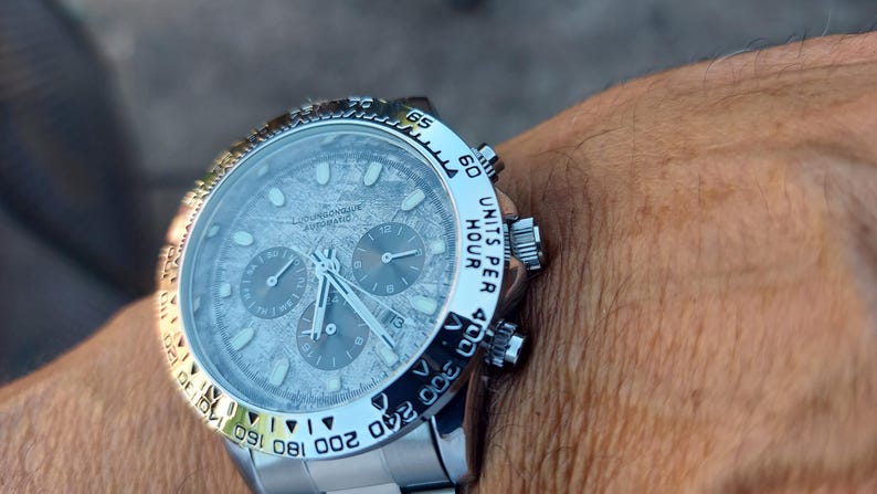 May include: A silver wristwatch with a textured gray face and three sub-dials. The watch has a silver bezel with black markings and a silver band. The watch face has the text "AUTOMATIC" and "UNITS PER HOUR".