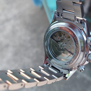 May include: A silver-toned wristwatch with a visible mechanical movement. The watch has a stainless steel band and a clear back, showcasing the internal gears. The watch is held at an angle, displaying its intricate design.