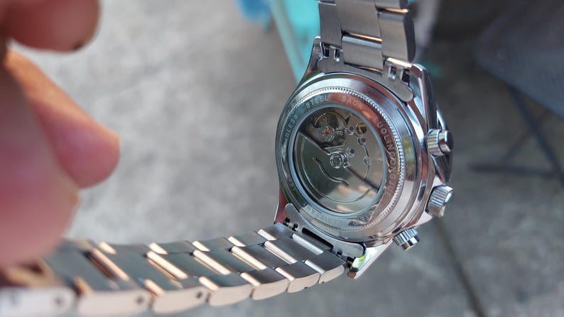May include: A silver-toned wristwatch with a visible mechanical movement. The watch has a stainless steel band and a clear back, showcasing the internal gears. The watch is held at an angle, displaying its intricate design.