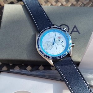 May include: A wristwatch with a blue and white face and a navy blue leather strap. The watch face has three sub-dials and the brand name "CILOA". The watch is displayed with its box and instruction manual.
