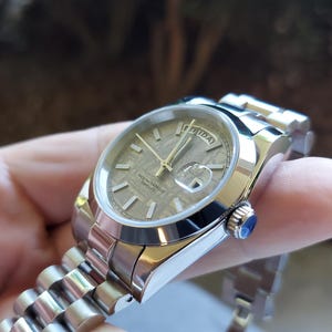 May include: A stainless steel wristwatch with a textured, light brown face and gold hands. The watch has a date display and a silver band. A small blue gem is set into the side of the watch.