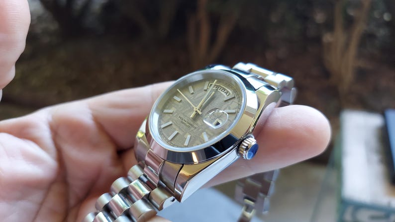 May include: A stainless steel wristwatch with a textured, light brown face and gold hands. The watch has a date display and a silver band. A small blue gem is set into the side of the watch.
