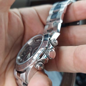 May include: A silver wristwatch with a black dial and a stainless steel strap. The watch features three small dials and a rotating bezel with numbers. The watch is held in a hand.