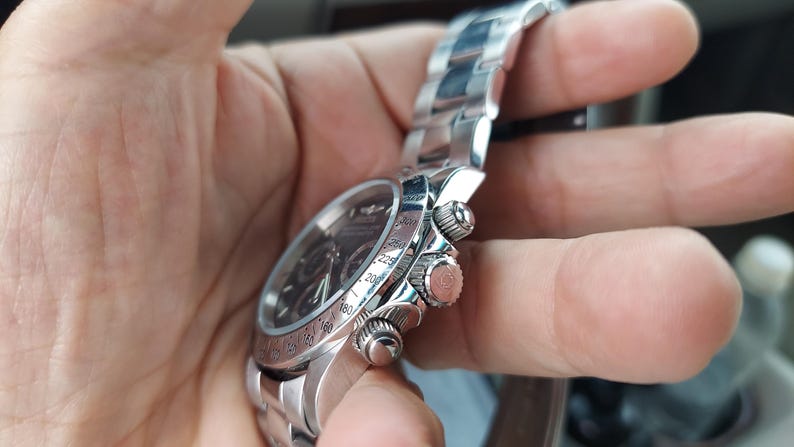 May include: A silver wristwatch with a black dial and a stainless steel strap. The watch features three small dials and a rotating bezel with numbers. The watch is held in a hand.