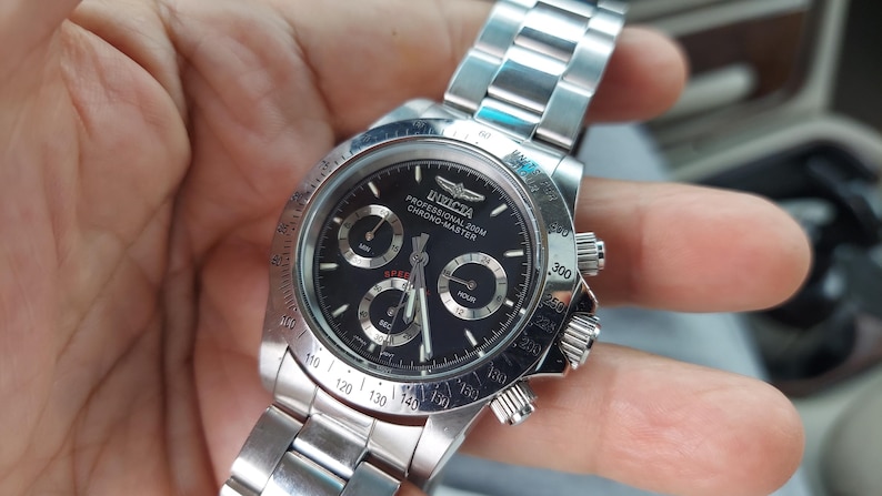 May include: A silver Invicta Professional 200M Chrono Master watch with a black dial. The watch features three sub-dials, a date display, and a stainless steel strap. The watch has a round face and a rotating bezel with numbers.