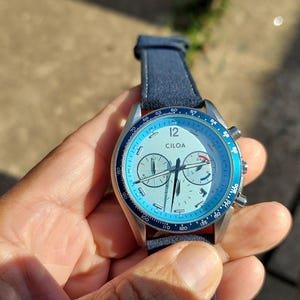 May include: A wristwatch with a light blue face and a dark blue leather strap. The watch face features the brand name "CILOA" and multiple sub-dials. The watch has a silver-colored metal casing and a blue bezel. The watch is being held in a hand.