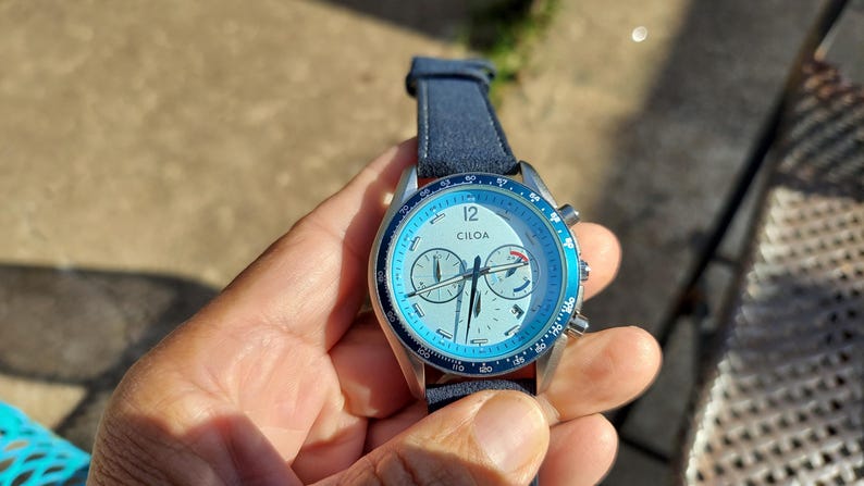 May include: A wristwatch with a light blue face and a dark blue leather strap. The watch face features the brand name "CILOA" and multiple sub-dials. The watch has a silver-colored metal casing and a blue bezel. The watch is being held in a hand.