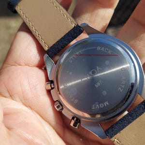 May include: A silver-toned watch with a light brown leather strap and navy blue accents. The watch face is visible, with the word "NOA" and "507" printed on it. The watch is a quartz movement.