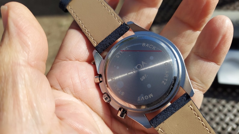 May include: A silver-toned watch with a light brown leather strap and navy blue accents. The watch face is visible, with the word "NOA" and "507" printed on it. The watch is a quartz movement.