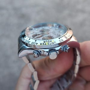 May include: A silver wristwatch with a light-colored face and a silver band. The watch has a bezel with black markings and three buttons on the side. The watch face displays multiple dials and hands.