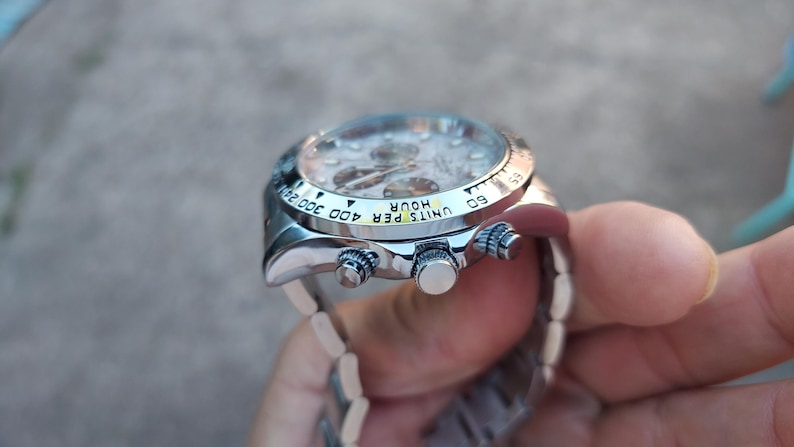 May include: A silver wristwatch with a light-colored face and a silver band. The watch has a bezel with black markings and three buttons on the side. The watch face displays multiple dials and hands.