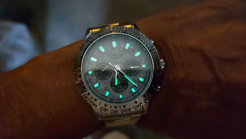 May include: A silver wristwatch with a dark gray face and luminous green markers and hands. The watch has three sub-dials and a rotating bezel with white numerals. The watch is on a silver band.