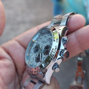 May include: A stainless steel wristwatch with a textured gray face and three sub-dials. The watch features a silver band and a bezel with black markings. The watch is held in a hand.