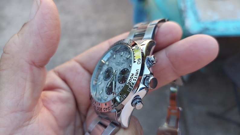 May include: A stainless steel wristwatch with a textured gray face and three sub-dials. The watch features a silver band and a bezel with black markings. The watch is held in a hand.