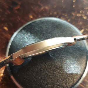 May include: A gold-toned wristwatch with a black leather band. The watch is resting on a dark, circular surface. The watch face is not visible, but the side profile shows a sleek, minimalist design.