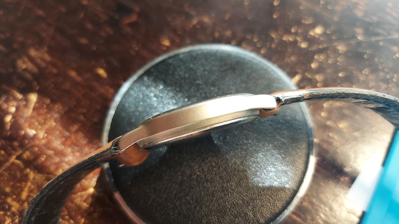 May include: A gold-toned wristwatch with a black leather band. The watch is resting on a dark, circular surface. The watch face is not visible, but the side profile shows a sleek, minimalist design.