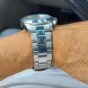 May include: A silver wristwatch with a stainless steel strap. The watch has a round face with a silver bezel and a gold-coloured crown. The watch is on a wrist, with the watch strap visible.