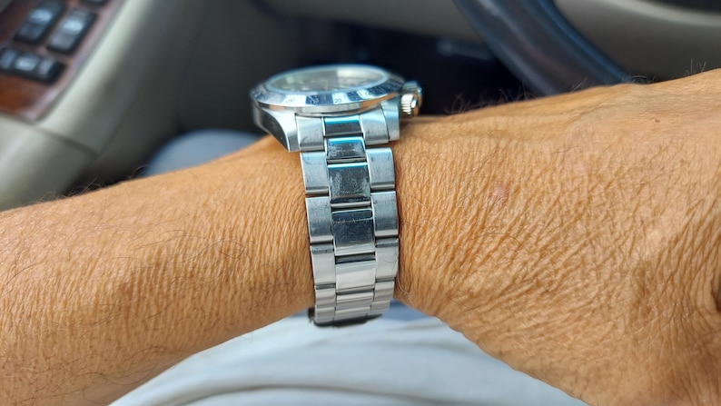 May include: A silver wristwatch with a stainless steel strap. The watch has a round face with a silver bezel and a gold-coloured crown. The watch is on a wrist, with the watch strap visible.