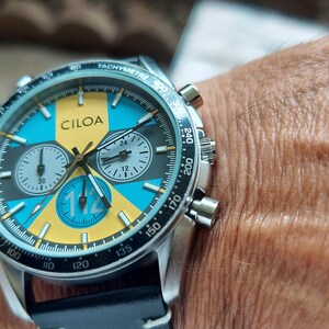 Pu&ograve; includere: Un orologio cronografo in acciaio inossidabile con cinturino in pelle nera. Il quadrante presenta un audace design geometrico in giallo brillante e blu turchese, con il marchio CILOA stampato in nero e il numero 112 ben visibile.