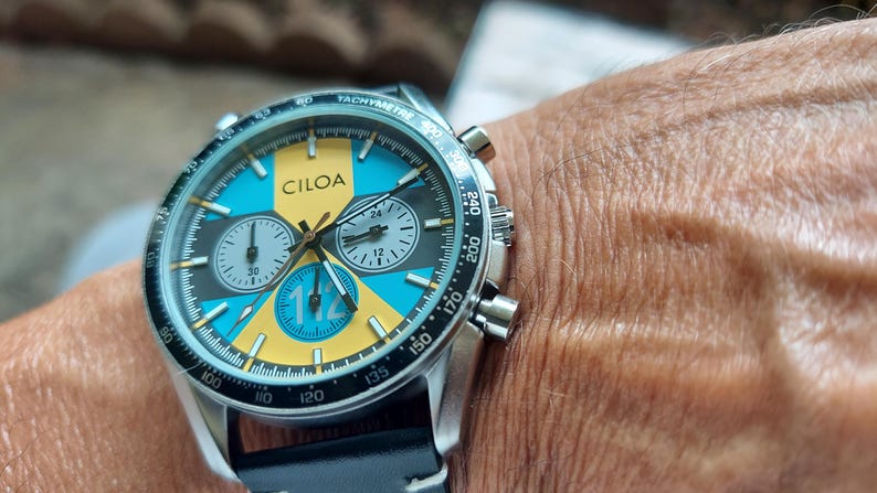 Pu&ograve; includere: Un orologio cronografo in acciaio inossidabile con cinturino in pelle nera. Il quadrante presenta un audace design geometrico in giallo brillante e blu turchese, con il marchio CILOA stampato in nero e il numero 112 ben visibile.
