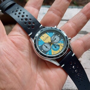 Pu&ograve; includere: Un orologio cronografo con cassa color argento e cinturino in pelle nera traforata. Il quadrante presenta un motivo geometrico in azzurro e giallo, con la scritta CILQA stampata in nero e tre sotto-quadranti di colore grigio.