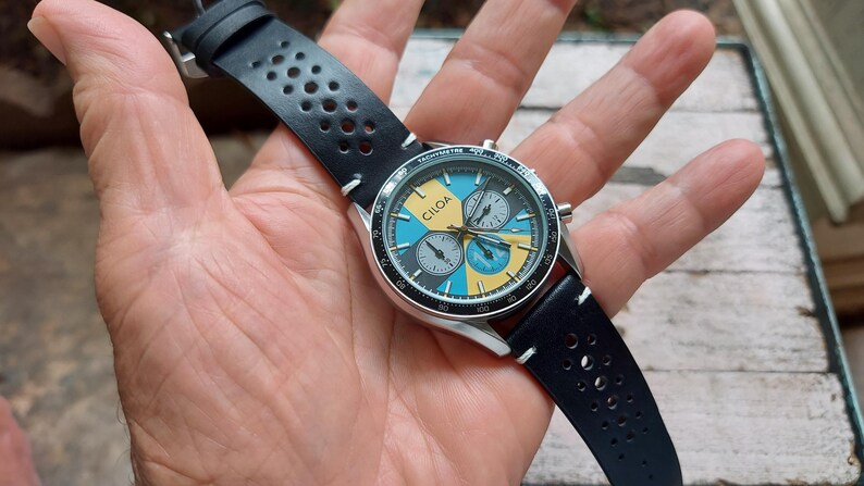 Pu&ograve; includere: Un orologio cronografo con cassa color argento e cinturino in pelle nera traforata. Il quadrante presenta un motivo geometrico in azzurro e giallo, con la scritta CILQA stampata in nero e tre sotto-quadranti di colore grigio.